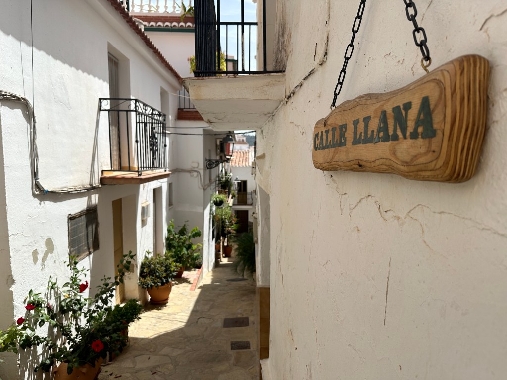 Zeer smal straatje met rechtss aan een witte muur een houten naambord: Calle Llana. 

