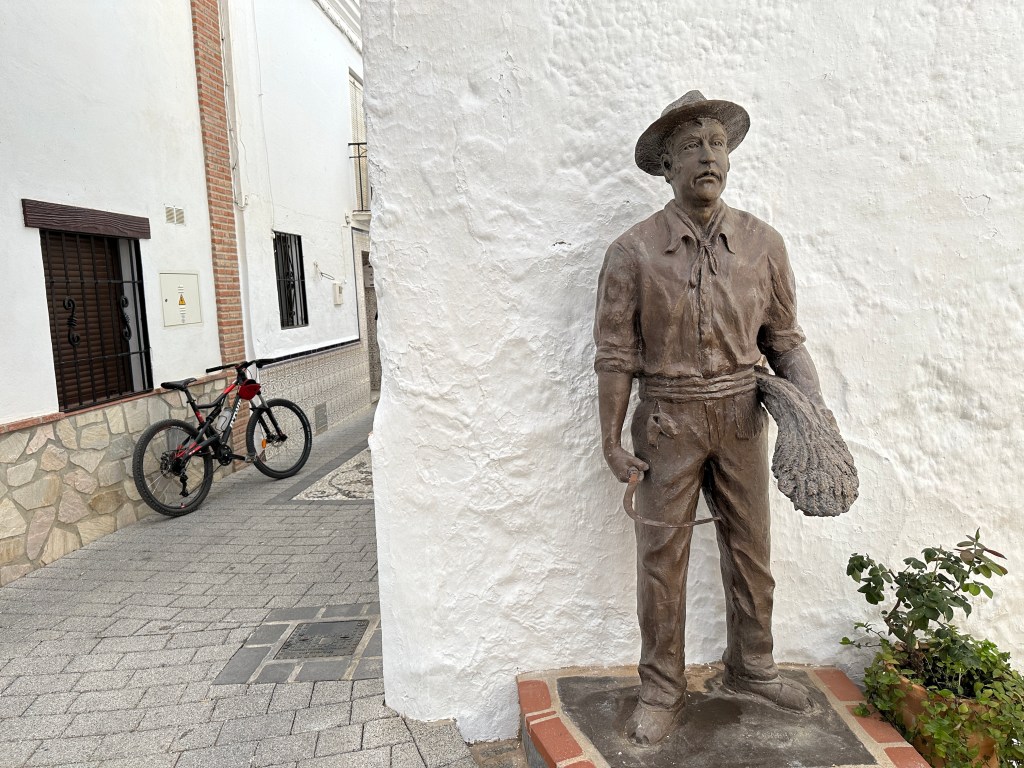Standbeeld van man met sikkel voor witte muur en zwarte fiets links. 