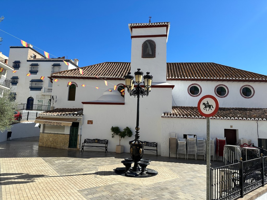 Dorpsplein van Arenas: wit gebouw met daarvoor lantaarnpaal met drie lampen. 