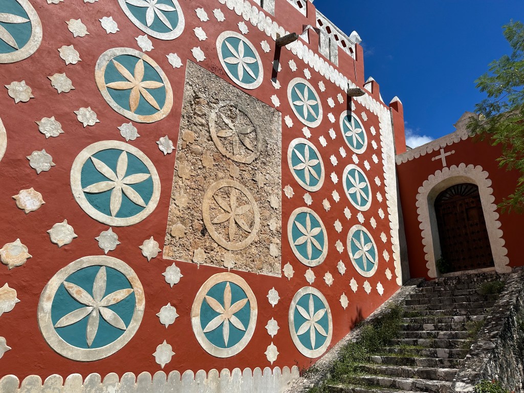 Gerestaureerde rode zijmuur van kerk met blauw-witte rozetten; twee ervan zijn niet gerestaureerd. 