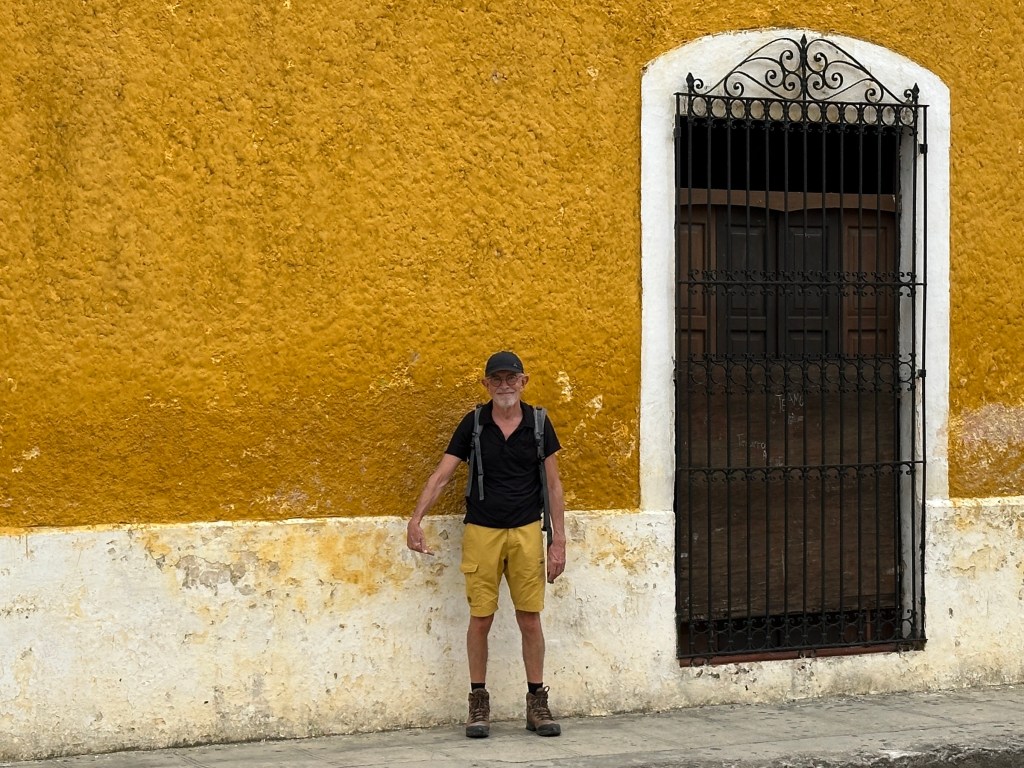 Toerist met gele broek voor okergeel huis met rechts zware, donkere deur. 