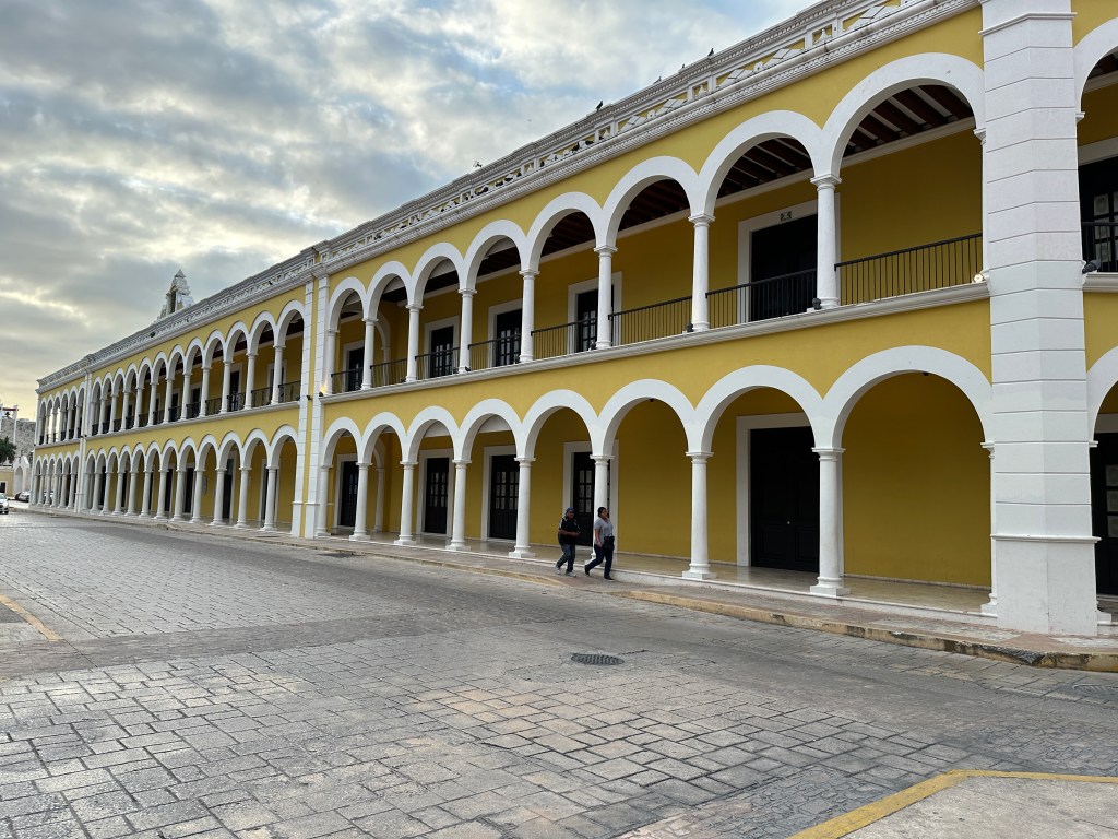 Lange straat en lang, geel gebouw met bogen op gelijkvloers en eerste verdieping in Campeche. 