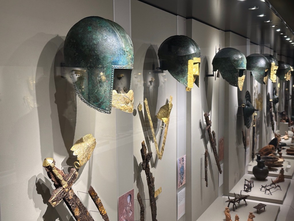 Tentoon gestelde rij helmen en gouden maskers in het archeologisch museum van Pella.