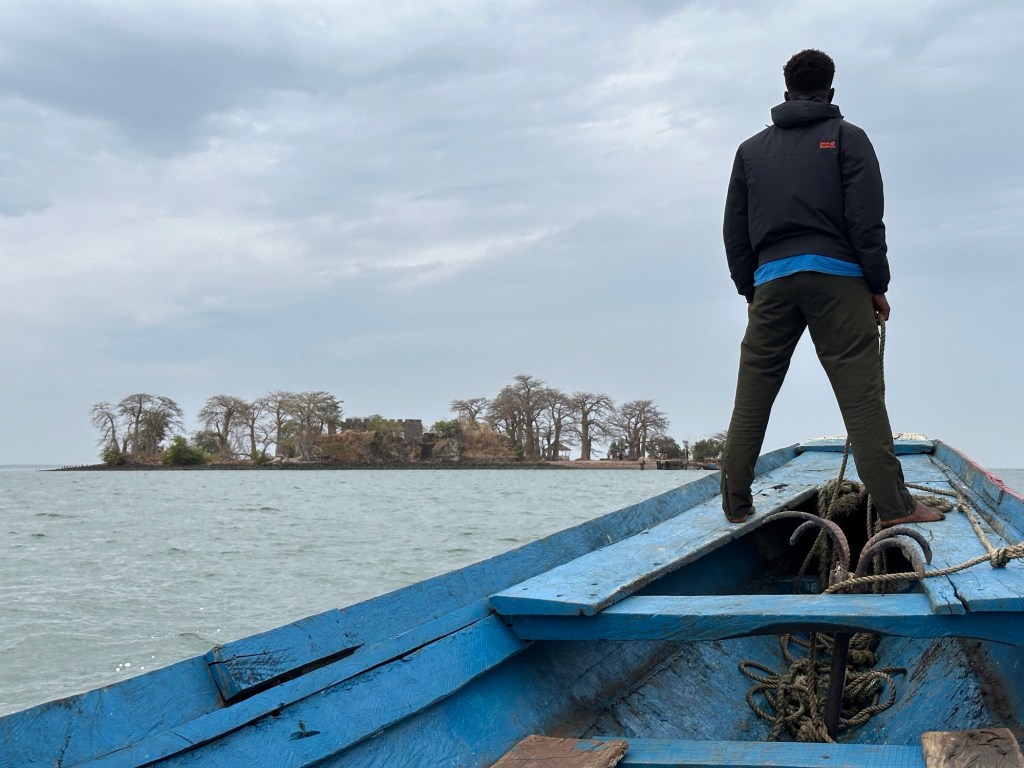 Voorsteven van boot met rechtopstaande man op Gambia rivier; Kunta Kinte eiland in de achtergrond
