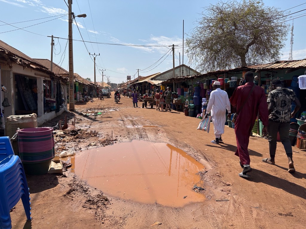 Straat van rode aarde met grote poel in het midden en wandelaars langs de kant