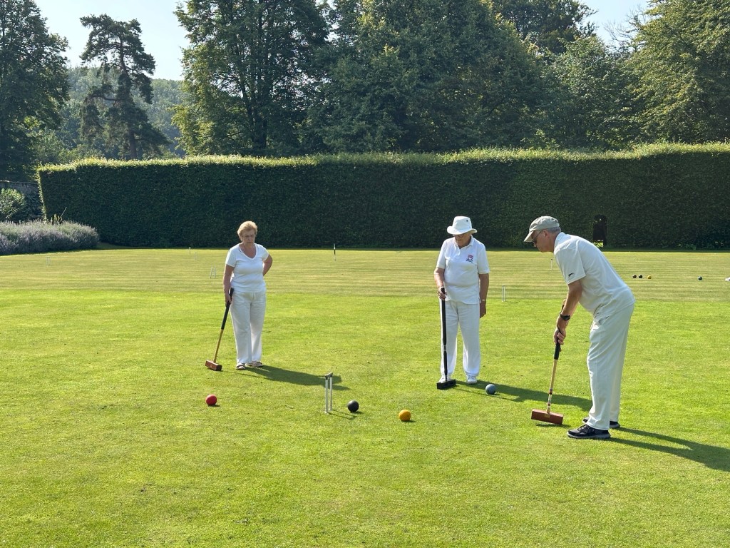 Drie spelers in witte kledij spelen croquet. 