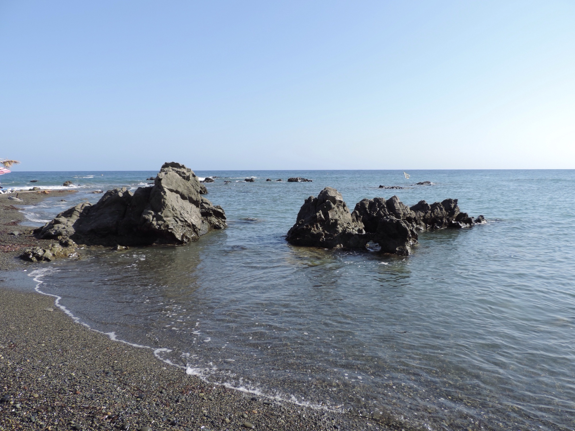 Grijs kiezelstrand met rotsen in blauwe zee.