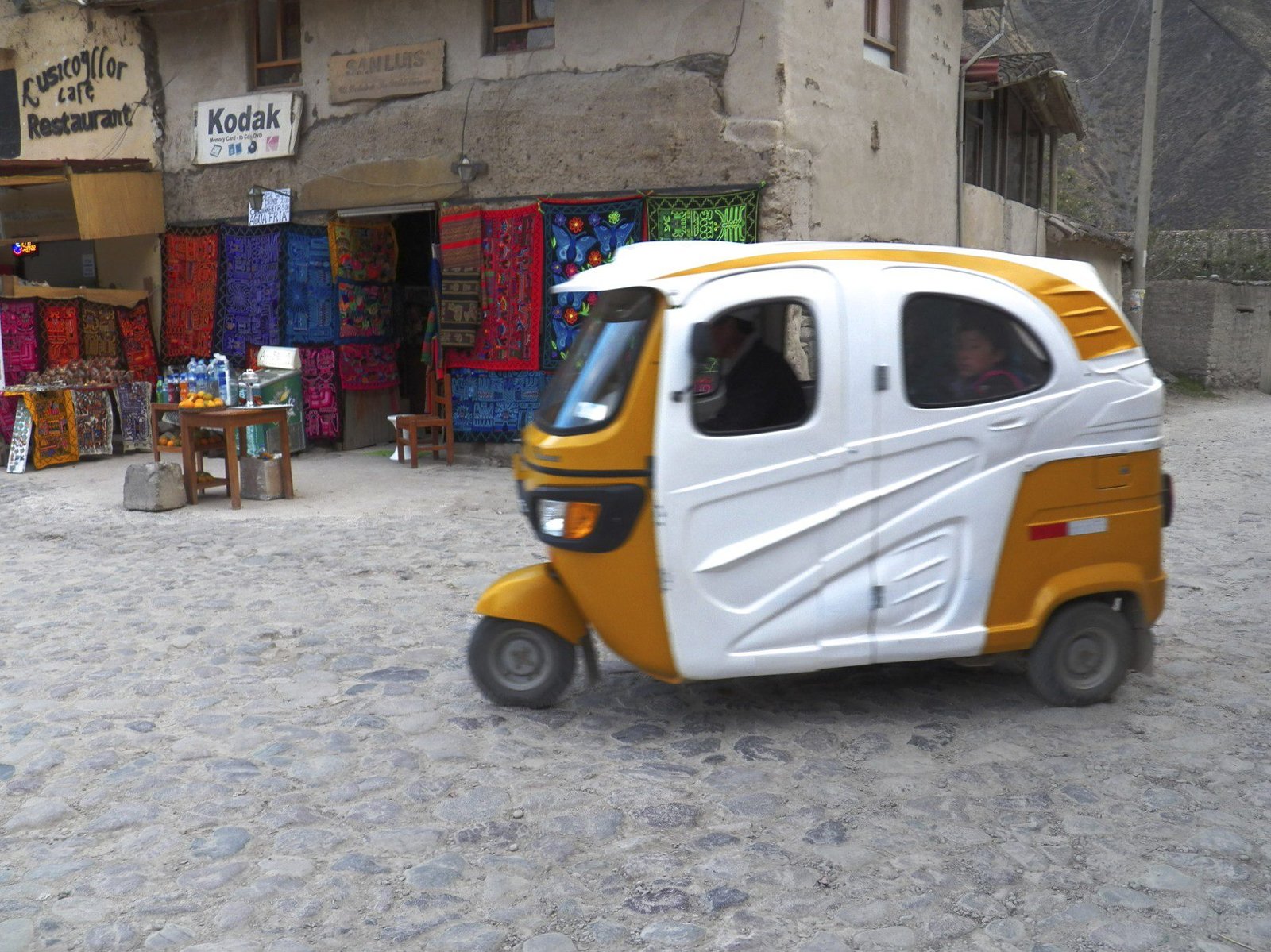 Geel-witte driewieler taxi voor winkeltje met kleurrijke tapijten en Kodak-reclame.