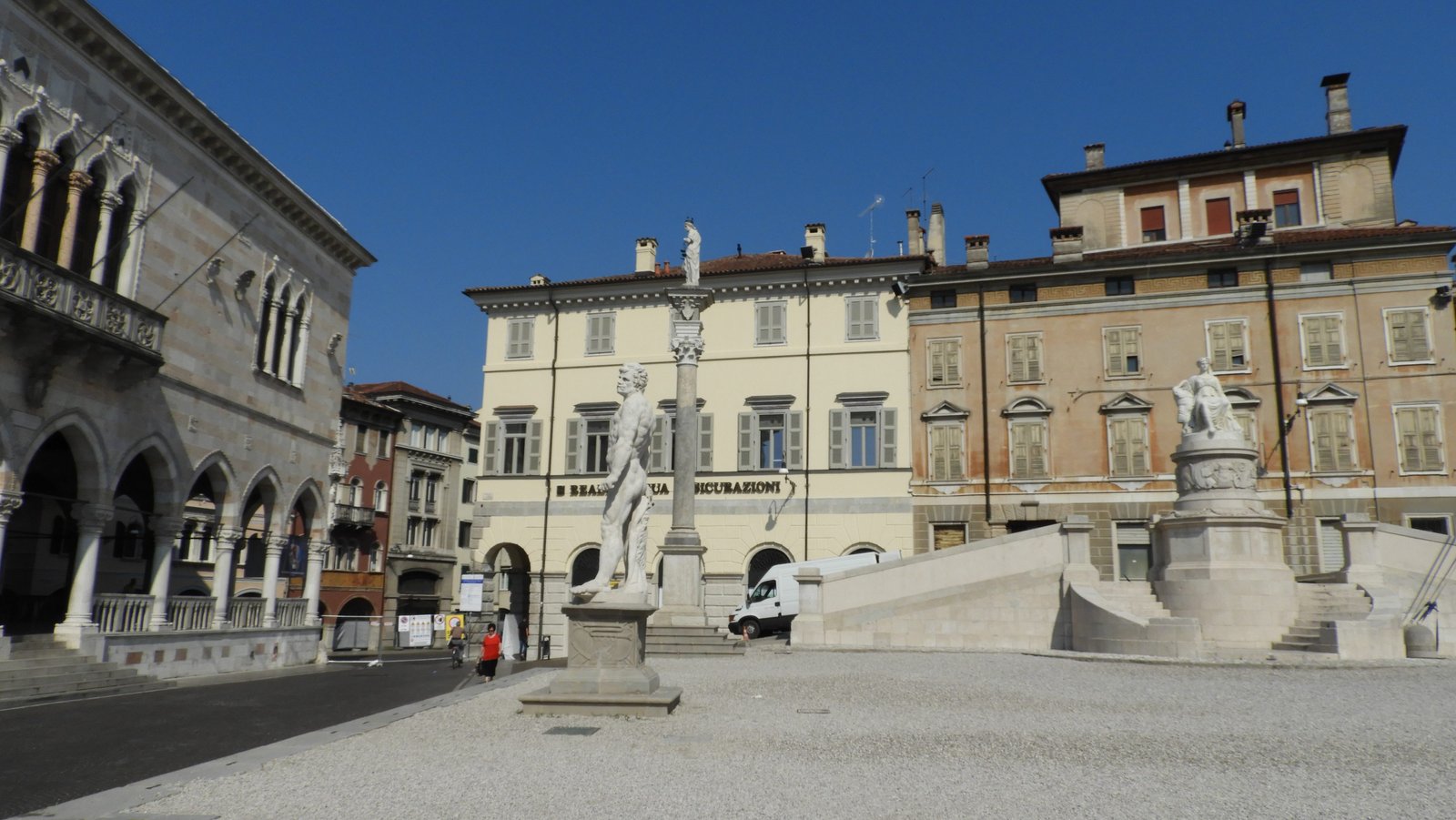 Plein in Udine met standbeeld links en beige en oker gebouwen.
