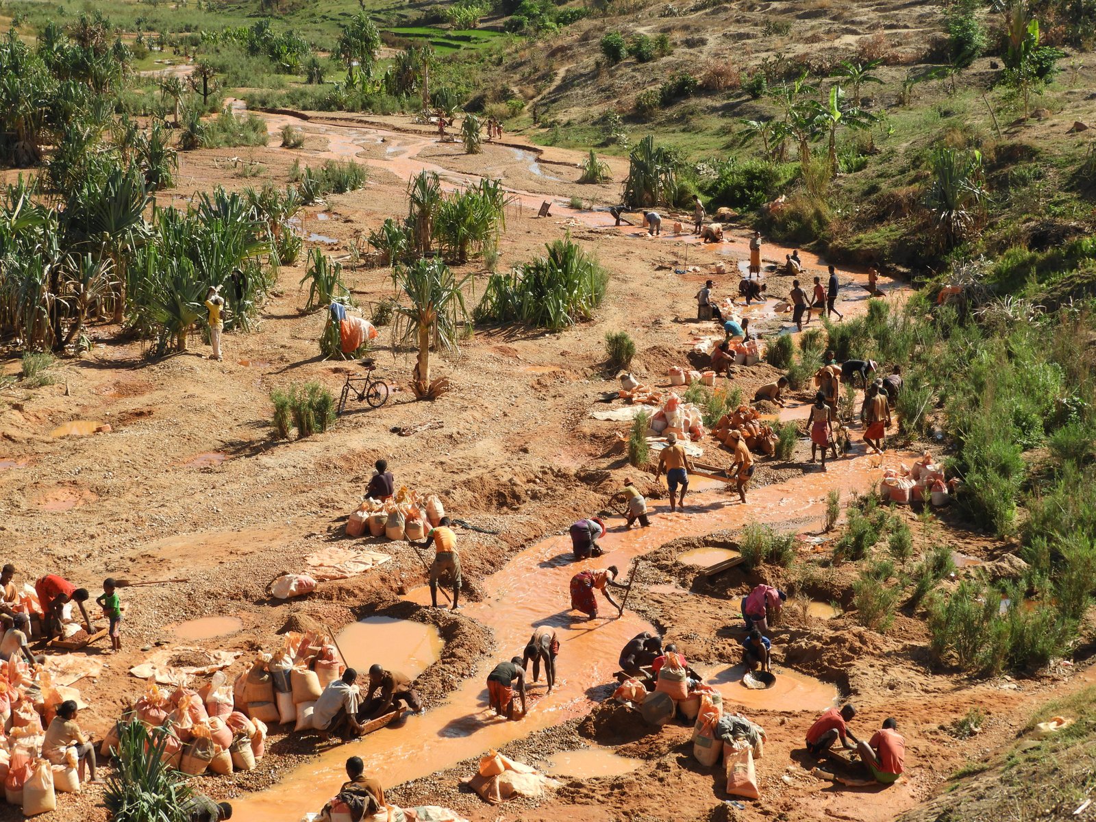 Mannen, vrouwen, kinderen aan een bruinrode rivier met modder en begroeiing.