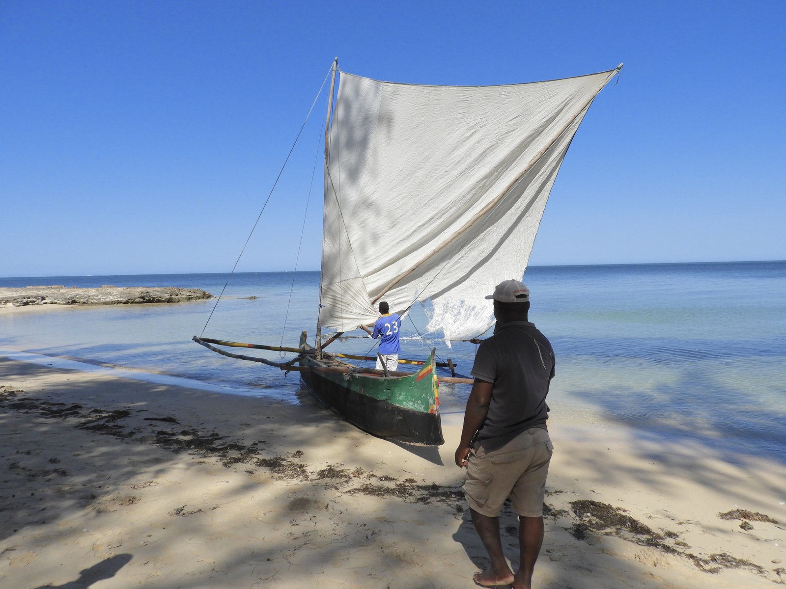Pirogue met groot wit zeil op rand van zee en strand.