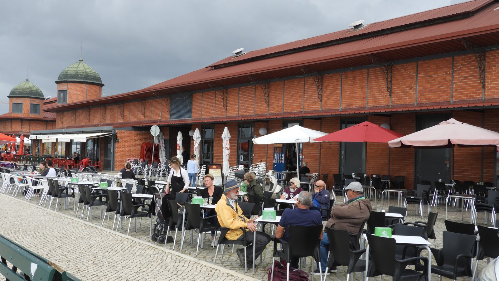 Lang rood gebouw met terrastafels en -stoelen er voor.