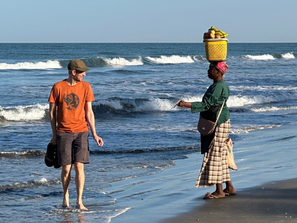Bumster (vrouw met gele mand op hoofd) spreekt een toerist met oranje T-shirt aan op het strand.