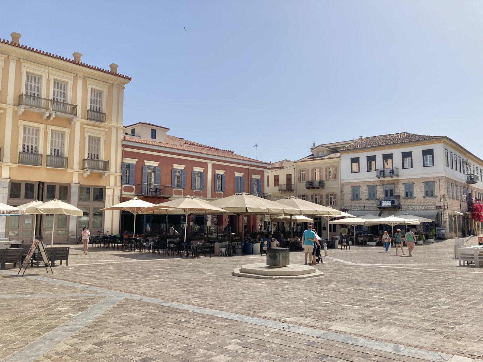 Plein in Nafplio met statige gele en oker-bruine huizen en beige parasols.