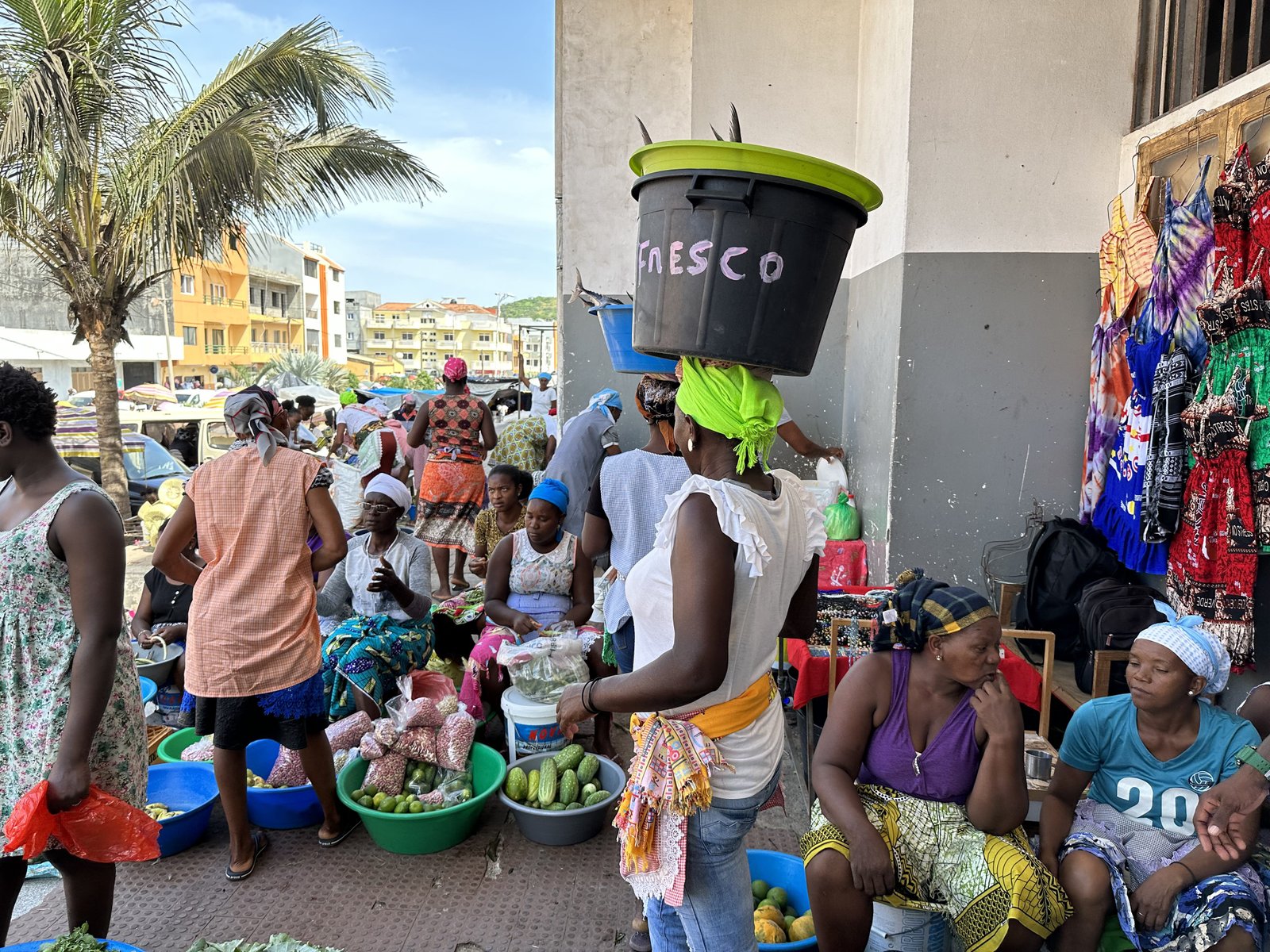 Kleurrijk geklede vrouwen waarvan twee met plastic kommen met vissen op het hoof op de Assomada markt.