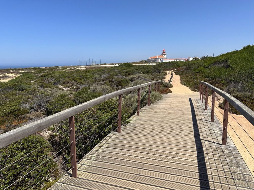Voetgangersbrugje op voorgrond, vuurtoren van Cabo Sardão in de verte