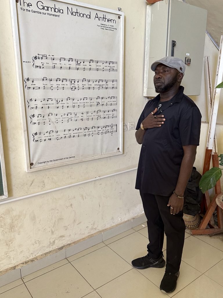 Gids zingt nationaal volkslied van Gambia naast affiche met tekst van volkslied