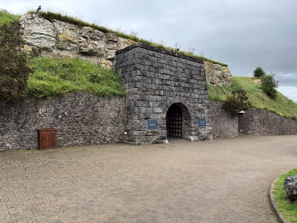 Muur in grijze steen met boog-ingang tot ondergronds gebouw. 
