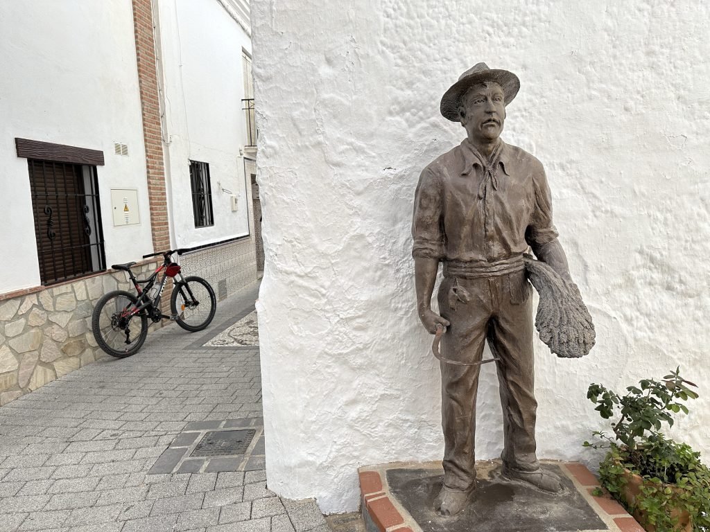 Standbeeld van man met sikkel voor witte muur en zwarte fiets links. 