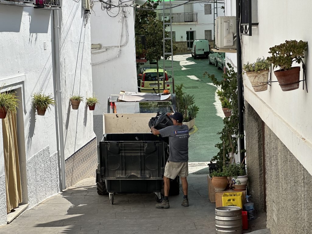 Vuilnisman laadt kleine vuilniswagen in smal straatje in Canillas de Aceituno. 
