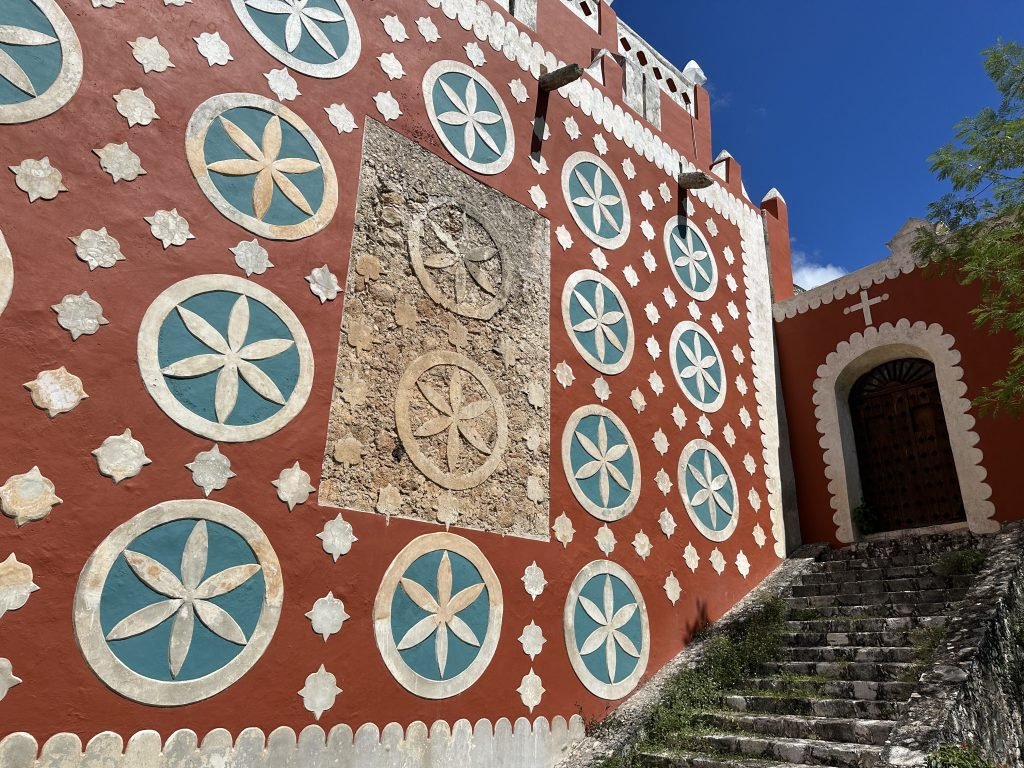Gerestaureerde rode zijmuur van kerk met blauw-witte rozetten; twee ervan zijn niet gerestaureerd. 