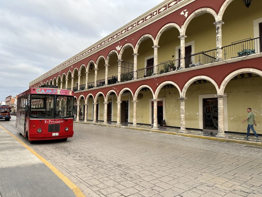 Lange straat en lang, rood gebouw met bogen op gelijkvloers en eerste verdieping; rode lijnbus links in Campeche. 